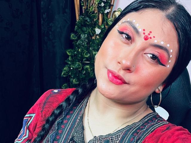 KalpanaAisha has bright red and pink eye makeup, white dots on her forehead, and red dots on her cheek. She's wearing a red patterned top and hoop earrings.