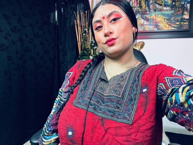 KalpanaAisha with medium tits, red face paint, braid, and red traditional dress, wearing colorful patterns, black hair, and hoop earrings, giving a slight pout.