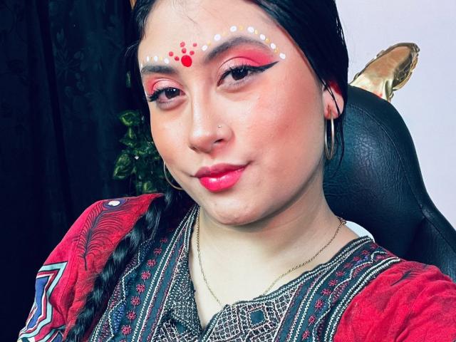 KalpanaAisha with dark hair, red lipstick, and white dot makeup on forehead, wearing red patterned top, black earrings, and gold necklace, sitting with black chair and golden statue.