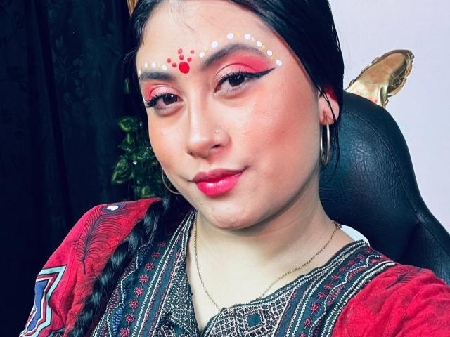 KalpanaAisha has red lipstick, pink eyeliner, white and red face paint, braided black hair, hoop earrings, nose ring, and wears a red patterned top with black and gold details.