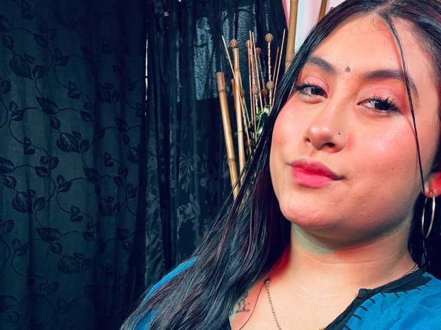 KalpanaAisha with long black hair, pink eyeshadow, red lipstick, and a nose ring, looking at the camera with a slight smile, wearing a blue shirt, showing her tits.