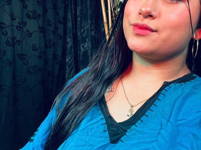 KalpanaAisha with long black hair, blue top, black trim, nose ring, and pink lips, showing a small tattoo on her right chest. She's wearing a gold necklace.