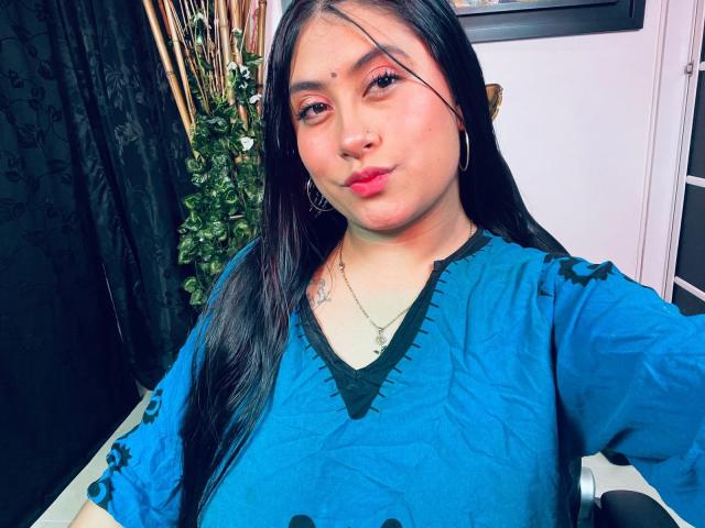 KalpanaAisha with long black hair, wearing a blue shirt, pouty lips, small nose ring, and gold necklace, has a tattoo on her right chest, looking at the camera.