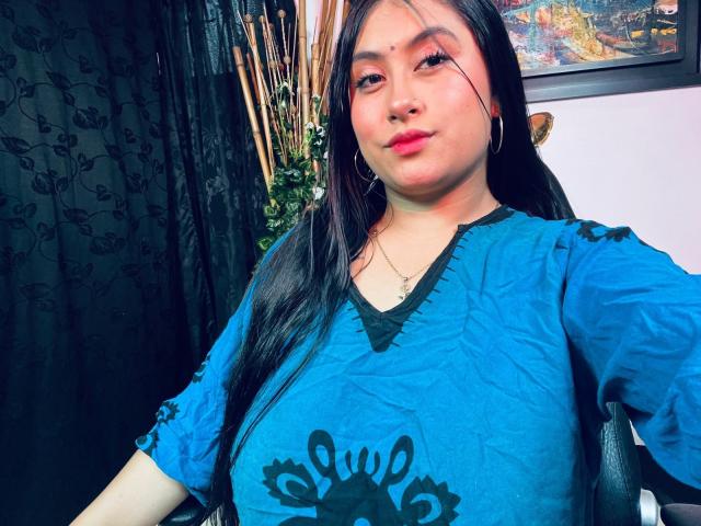 KalpanaAisha has big tits in a blue shirt with black patterns, long black hair, red lipstick, and hoop earrings, sitting in a chair, looking at the camera.