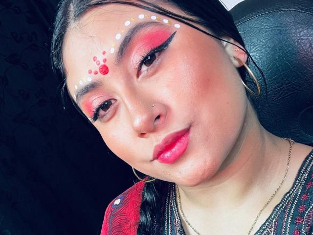 KalpanaAisha with bright red eyeshadow, white dot face paint, pink lipstick, and small nose ring, wearing hoop earrings and a red patterned top, looking slightly to the side.