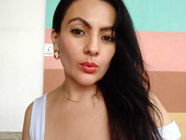 JakyKidman has dark hair, brown eyes, red lipstick, and wears a white top with small tits, a necklace, and hoop earrings. She's making a pouty face.