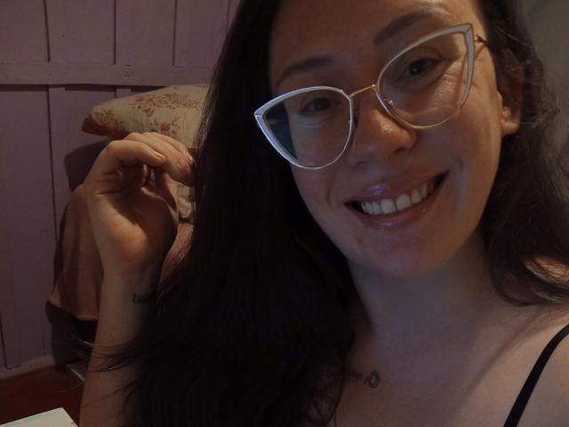 LunnaBellamore with glasses, smiling, dark hair, small tits, tattoo on right wrist, pinkish skin, shiny lips, holding hair, pink wall in background, cozy indoor setting, looking cute.