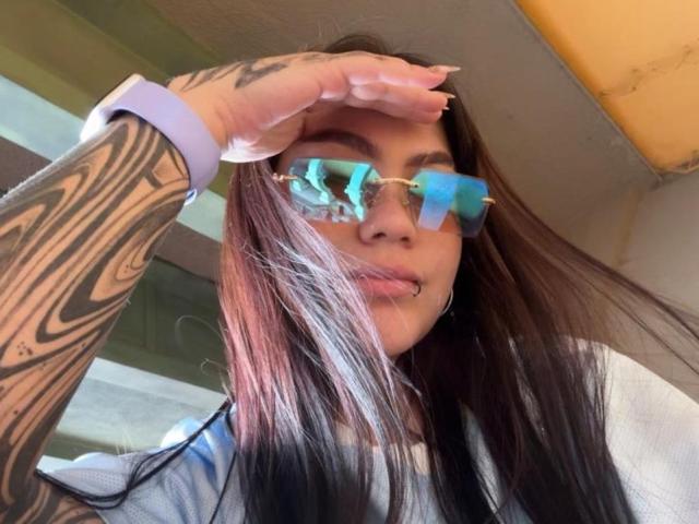 TessaMagic has long brown hair, reflective blue sunglasses, and a tattooed arm, sticking out her tongue. She's wearing a white top and a purple wristband.