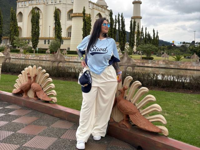 TessaMagic stands in front of a detailed, carved stone bench with turkey shapes, wearing a light blue "Said" shirt, white pants, and white sneakers. She has long dark hair, blue glasses, and a navy handbag. The background features a large, ornate building with multiple towers and a lush, green landscape. The stone bench is positioned on a checkered pavement. The image is clear with a focus on TessaMagic's relaxed, confident posture.