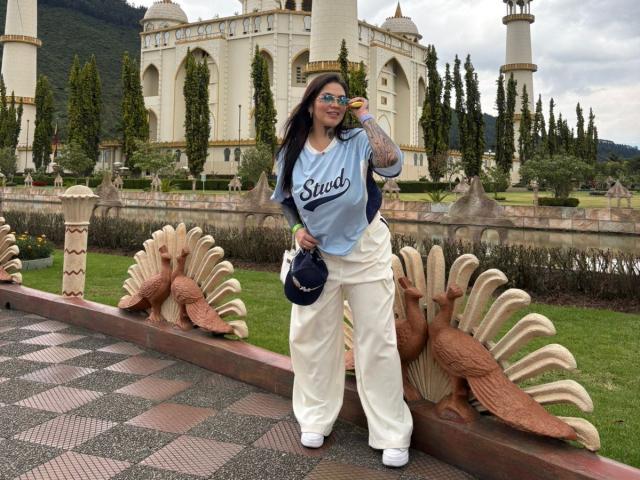 TessaMagic stands in front of a detailed, ornate building with multiple domes and columns. She wears a light blue "Sandy" t-shirt, white pants, and white sneakers. She holds a black purse in her right hand and eats a donut with her left. Her long dark hair flows over her shoulders. The building features carved bird motifs and a checkered path. The image has a natural, overcast light.