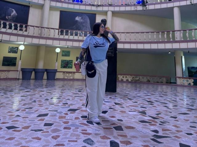 TessaMagic with tattoos, blue shirt, white pants, and high heels, sipping from a red cup, standing in a shiny, patterned tiled room with a balcony.