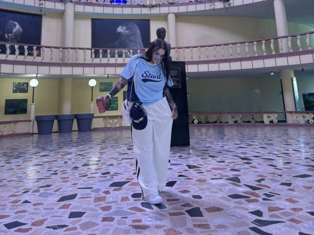 TessaMagic stands in a multi-level indoor space with a mosaic-tiled floor. She wears a light blue "Steee" t-shirt, loose white pants, and white sneakers. She holds a red drink in her right hand and a black purse in her left. She has tattoos on her right arm and is wearing sunglasses. The background features a balcony with decorative railings and large paintings of animals. The lighting is even, and the space is well-lit. The composition centers on TessaMagic, with her slightly leaning forward. The mosaic tiles are varied in color, including black, white, and brown.