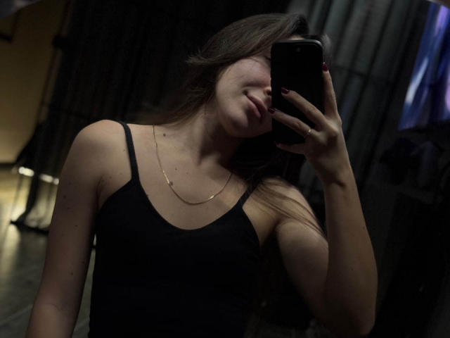 AquaLady with long brown hair, wearing a black tank top, holding a phone in front of her face, showing her tits and a gold necklace, in a dimly lit room.