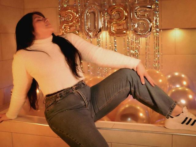 PaulinaCox with long black hair, white turtleneck, blue jeans, and white Adidas sneakers, leaning back with one leg raised, showing her ass. Gold balloons and 2015 balloons in background.