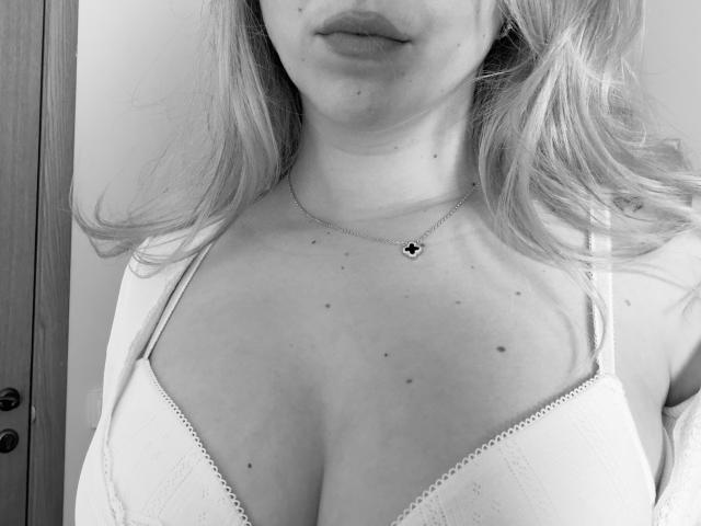 SugarrX's upper chest with small, dark moles, wearing a white bra with lace trim, a delicate necklace, and loose, wavy blonde hair. Boobs are slightly visible.