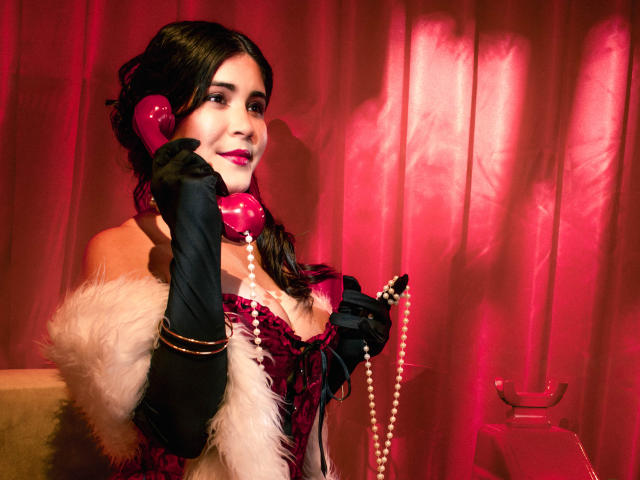 EvaSingh with dark hair, pink phone, black gloves, white fur, and pearl necklace, smiling, holding phone to ear, red background, pink lighting, pink lips, cleavage, seductive look.