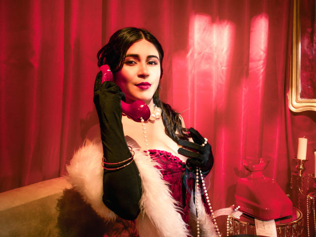 EvaSingh with red lipstick, black gloves, and white fur, holding pink phone, wearing pink and black corset, pearl necklace, and pink gloves, in red-lit room.