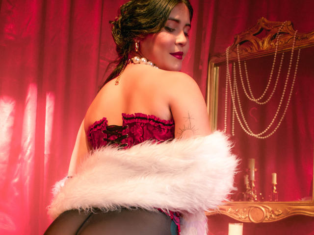 EvaSingh with red velvet corset, white fur, black panties, pearl necklace, tattoo on left arm, dark green hair, red lipstick, looking back, red curtain, gold mirror, pearl necklace.