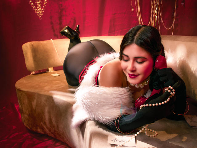 EvaSingh lies on a beige couch, wearing black gloves, a red and white fur-trimmed outfit, and black heels, holding a pearl necklace, with her ass and tits visible.