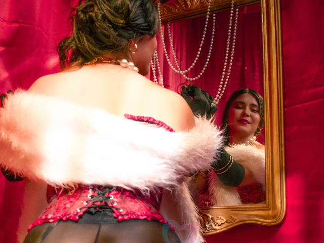 EvaSingh in red lace corset, black gloves, and pink fur, facing mirror, reflecting her dark hair and red lipstick, pearl necklace, and gold frame, against pink curtain.