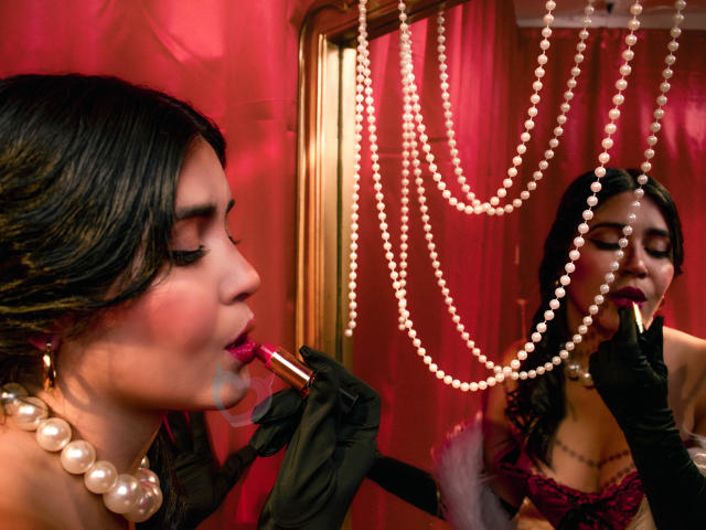 EvaSingh with dark hair, pearl necklace, black gloves, and pink lipstick, applying makeup in a mirror, wearing a red bra, pearl necklace reflected, red curtains in background.