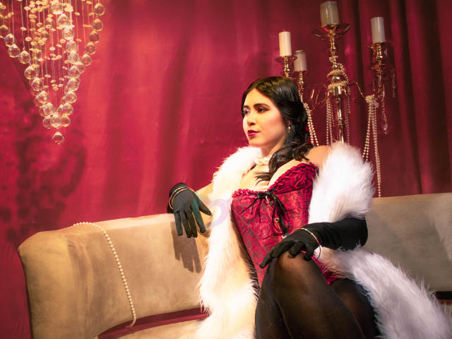 EvaSingh with black hair, red corset, black gloves, white fur coat, sitting on beige sofa, red background, candelabras, looking to the side, showing her ass.