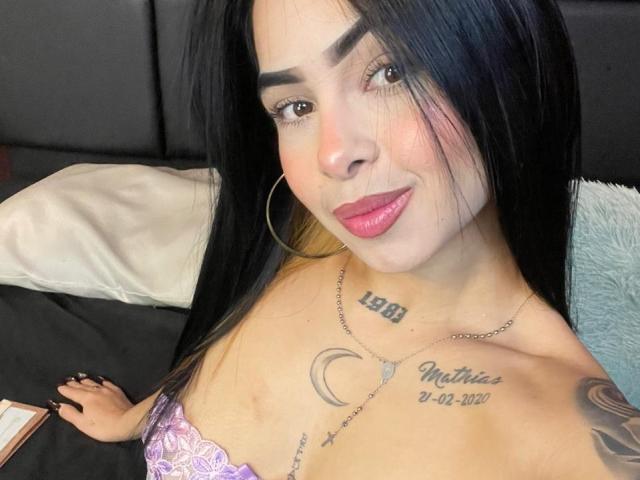 MistressAsshley ha lunghi capelli neri, rossetto rosa, orecchini a cerchio e tatuaggi, tra cui "LEBI" e "Chantias" sul petto. Sta sorridendo, mostrando il seno in un reggiseno di pizzo viola..