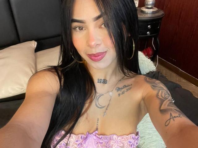 MistressAsshley with long black hair, hoop earrings, pink lipstick, tattoos on chest and arm, purple strapless top, showing her tits, smiling at the camera.