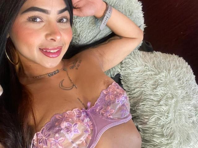 MistressAshley, a Latina woman with long black hair, smiles at the camera. She has dark eyebrows, pink lipstick, and light brown skin. She wears a pink and purple lace bra with floral patterns, exposing her small, brown areolas. Her right arm, with a grey wristwatch, rests above her head. She has a crescent moon tattoo on her right chest and a tattoo on her left shoulder. She's lying on a fluffy green pillow. The composition is a close-up, focusing on her upper body.