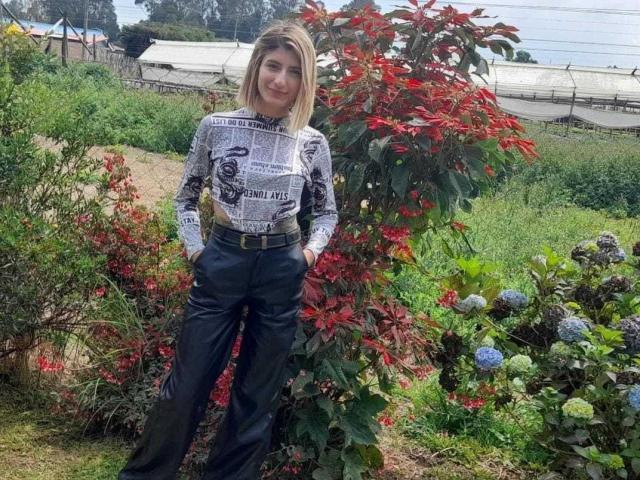 Kaiix standing in a garden with red leaves, wearing a newspaper-print shirt and black pants, hands in pockets, smiling, blonde hair, green foliage, blue flowers, sunny day.
