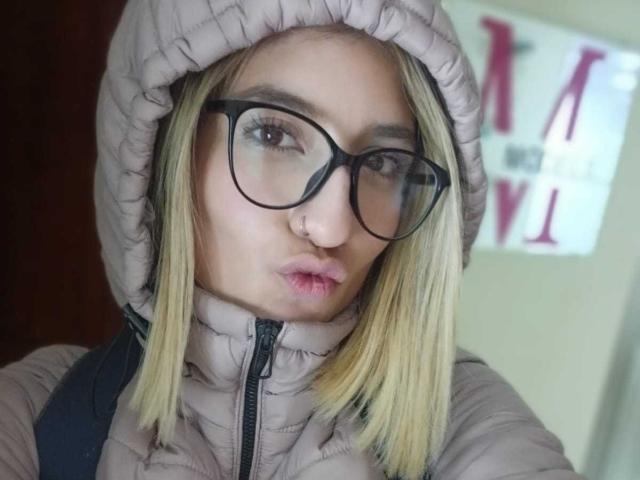 Kaiix has straight, shoulder-length blonde hair, black-framed glasses, and a nose ring. She wears a puffy, beige jacket with a quilted hood. Her lips are slightly puckered. The background is blurred, featuring a white sign with red text. The image has a natural, slightly overexposed look. Kaiix's glasses have a thick, modern style. Her expression is neutral, and the photo is a close-up, focusing on her face and upper jacket.