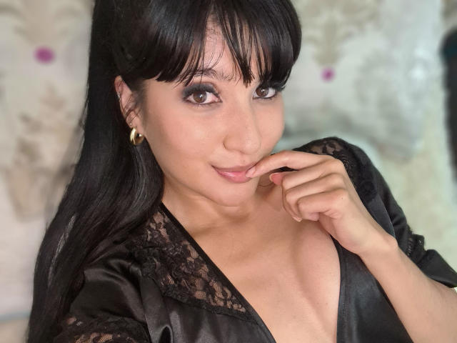 NaylaGrey vestindo uma camisa de renda preta, com o dedo nos lábios rosados, sorrindo para a câmera, usando brincos de argola dourados, olhos castanhos, cabelo preto longo, pele clara, mostrando o decote..
