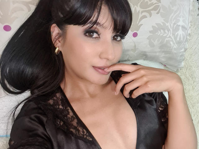 NaylaGrey with black hair, wearing a black lace top, biting her finger, showing off her tits, and looking sexy with gold earrings. She's lying on a fluffy pillow.