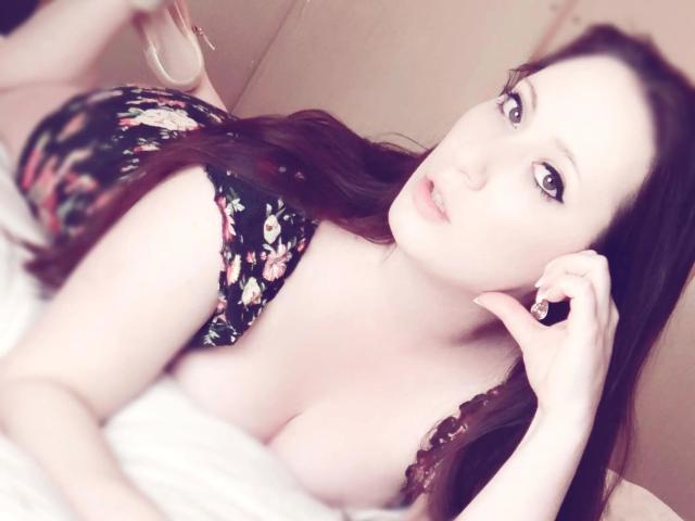 LaviniaJolie-hot with pale skin, brown hair, and blue eyes, lying on a bed in a floral dress, showing her big tits, and looking at the camera with a slight smile.