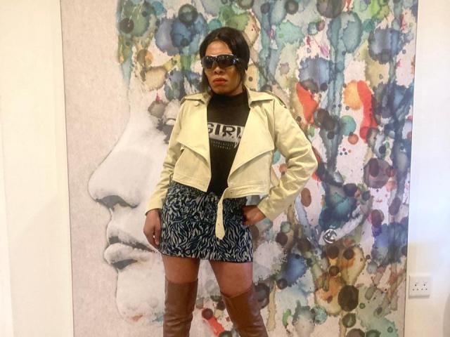 ImaniJohnson stands in front of abstract art, wearing a yellow leather jacket, black turtleneck, blue patterned skirt, and brown thigh-high boots, with black sunglasses and a confident pose.