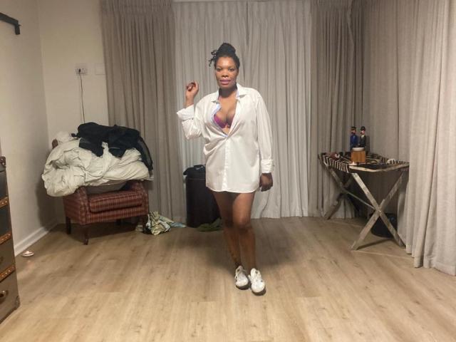 ImaniJohnson, dark-skinned woman with curly hair, wearing a white shirt unbuttoned to show pink bra, white sneakers, standing in room with wooden floor, smiling, holding shirt edge.