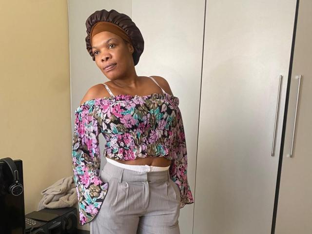 ImaniJohnson with brown skin, wearing a floral off-shoulder top, gray pants, and brown headwrap, standing in a room, showing her midriff and white underwear.