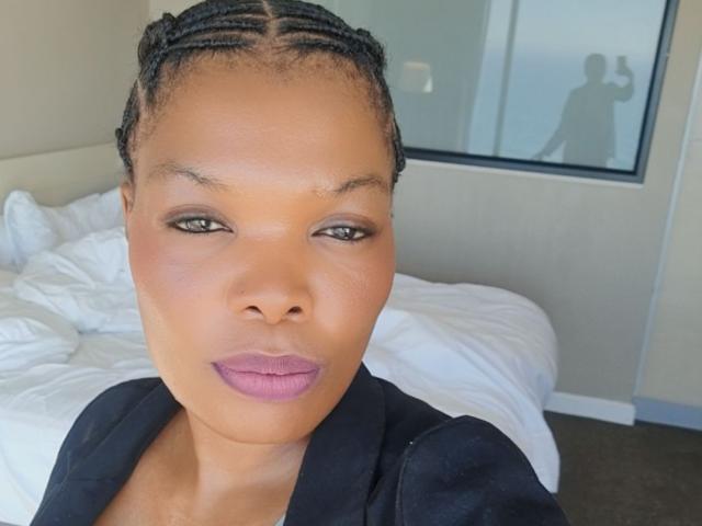 ImaniJohnson with braided hair, purple lipstick, and black jacket, standing in a room with a messy bed. Her ass is not visible. Her face is centered in the photo.