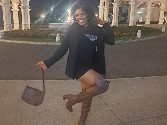 ImaniJohnson is smiling with her mouth open, wearing a black hoodie, short skirt, and brown boots, holding a brown purse, and has a dark skin tone, curly hair.