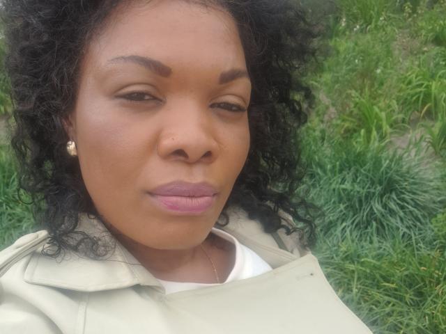 ImaniJohnson has dark curly hair, brown skin, purple lipstick, a nose piercing, and wears a beige jacket with small white earrings. She's looking slightly to the side.