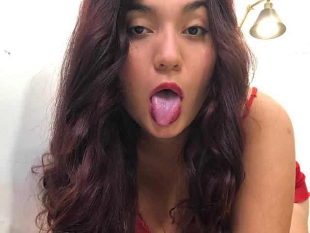 IvyJoness met lang, golvend, donkerpaars haar, rode lippenstift en haar tong uit, draagt rode lingerie, onder een zilveren lamp. Haar ogen zijn half open en ze kijkt naar de camera..