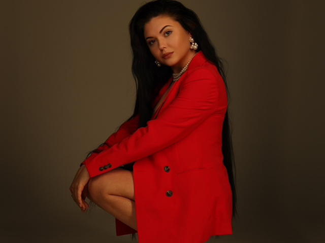 XKleo with long black hair, red blazer, pearl necklace, and earrings, squatting, showing one knee, looking at camera, dark brown background. Boobs, ass, and tits visible.