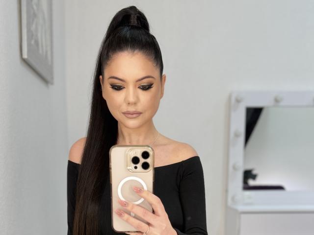 XKleo with long black ponytail, off-shoulder black top, holding gold phone, looking at screen, mirror with bulbs in background, eyeliner, nude lipstick, serious expression, elegant.