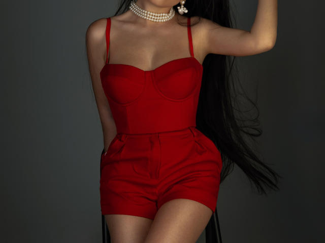 XKleo with long black hair, wearing a red strapless bustier and matching high-waisted shorts, a pearl choker, and a diamond earring, holding her arm up.