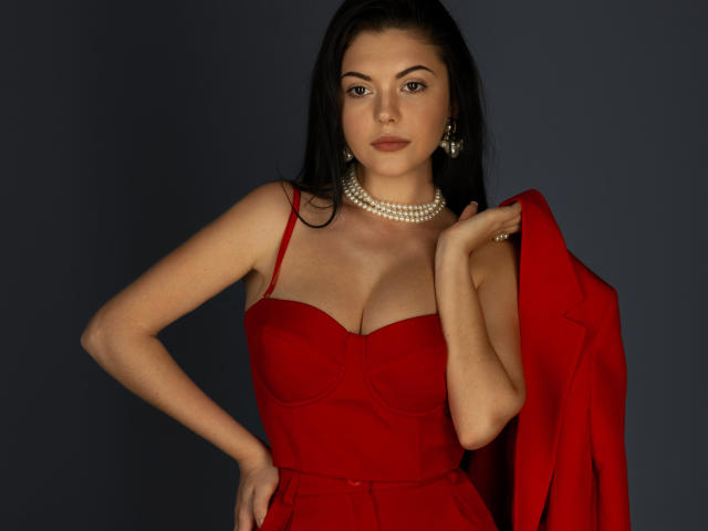 XKleo with red dress, pearl necklace, and black hair, holding a red jacket, showing her tits, looking confident, dark background.