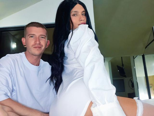CoupleSensualHot with long black hair, white shirt, and white panties, sits on bed with white shirted man, showing her ass, looking back, in bright room.
