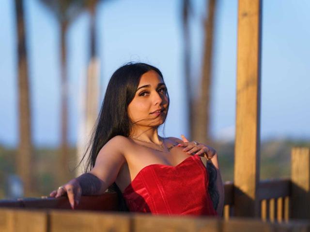 DaniXvg with long black hair, wearing a red strapless dress, showing small tits, tattoo on right arm, sitting on wooden balcony, hands on chair, blue sky in background.
