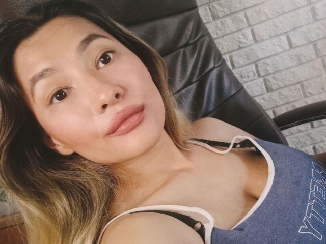 DamelySakura with her puffy tits in a blue tank top, lying back in a black chair, looking at the camera with a subtle smile. Her lips are slightly parted.