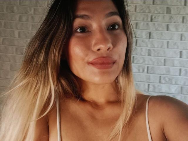 DamelySakura with a cute smile, light brown skin, and blonde ombre hair, wearing a white spaghetti strap top, showing her tits and puffy ass in the selfie.