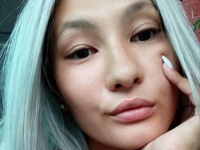 DamelySakura's face with pale blue hair, brown eyes, and white lipstick. She's touching her cheek with a white-nailled hand, showing a subtle smile. Her nose is small.
